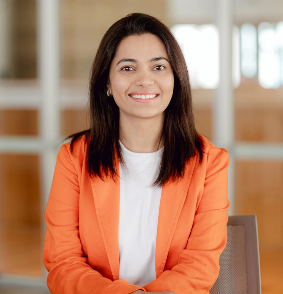Photo of a woman with black hair wearing an orange jacket with a white top underneath.