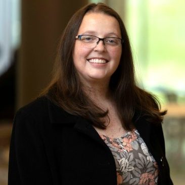 The image shows a professional portrait of a woman with long, dark brown hair and glasses, smiling warmly. She is wearing a floral-patterned blouse under a black jacket, and the setting is softly lit, with an out-of-focus background featuring natural light and hints of greenery. The photograph conveys a friendly and approachable demeanour, suitable for professional or academic contexts.